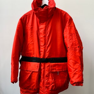May include: A bright red, long-sleeved, puffer jacket with a black belt and a zipped pocket on the front. The jacket has a high collar and a drawstring at the bottom.