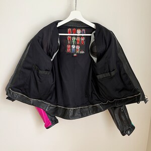 May include: Black leather motorcycle jacket with pink and teal accents. The jacket is unzipped and has a mesh lining. A label with a graphic of racing suits is visible on the inside of the jacket.