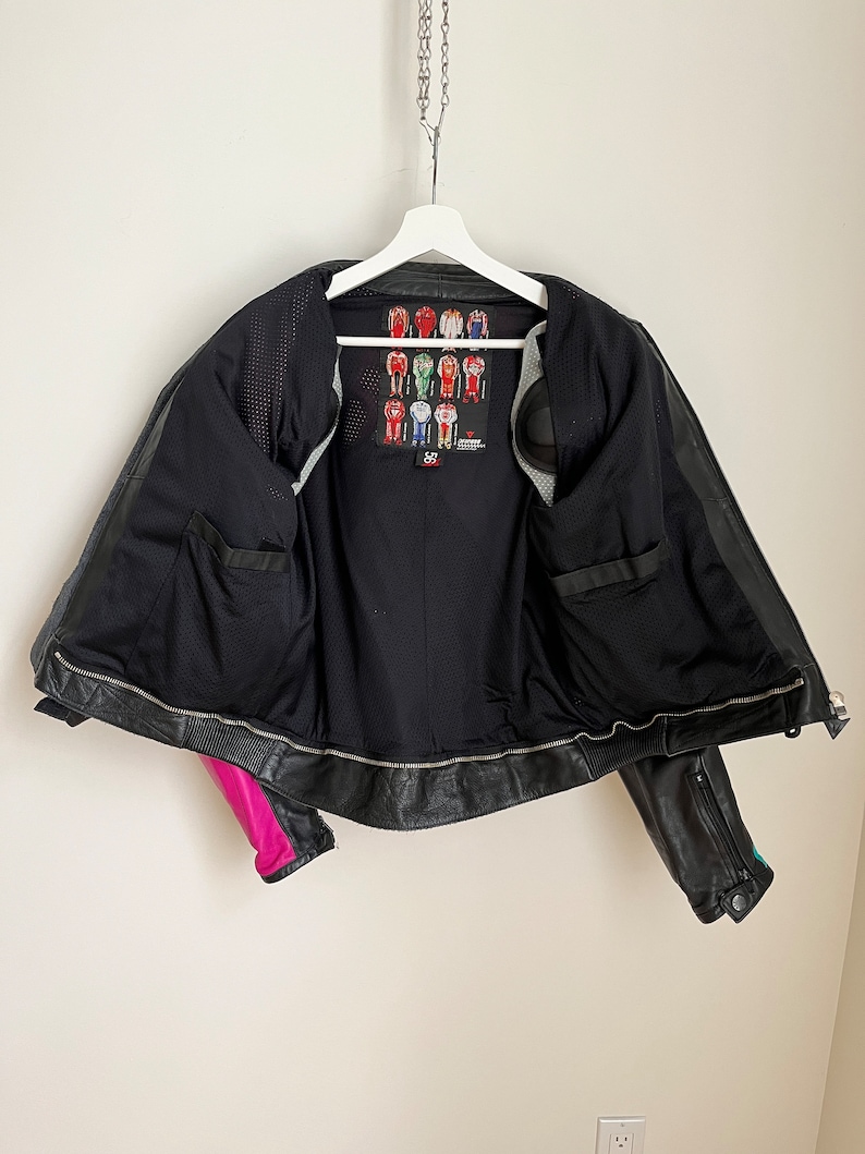 May include: Black leather motorcycle jacket with pink and teal accents. The jacket is unzipped and has a mesh lining. A label with a graphic of racing suits is visible on the inside of the jacket.