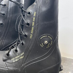 May include: A pair of black rubber boots with a fold-down gusset and a valve on the side. The boots have a yellow label that reads "KEEP DOUBLE LACED TO HOLD FIRMLY" and "FOLD GUSSET THIS SIDE ONLY". The valve has a label that reads "UNLESS AIRBORNE KEEP CLOSED OPEN".
