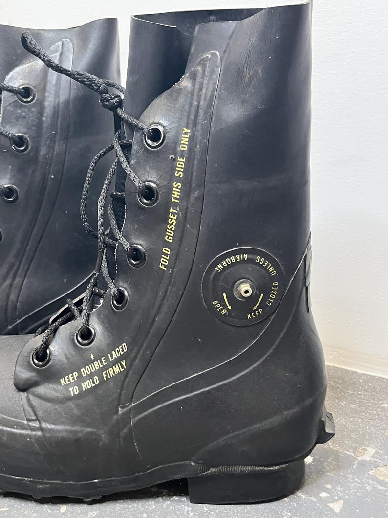 May include: A pair of black rubber boots with a fold-down gusset and a valve on the side. The boots have a yellow label that reads "KEEP DOUBLE LACED TO HOLD FIRMLY" and "FOLD GUSSET THIS SIDE ONLY". The valve has a label that reads "UNLESS AIRBORNE KEEP CLOSED OPEN".