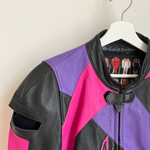 May include: A black, pink, and purple leather motorcycle jacket with a zipper closure. The jacket has a  distinctive design with geometric shapes and a  triangle logo on the front.