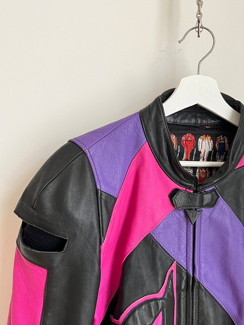 May include: A black, pink, and purple leather motorcycle jacket with a zipper closure. The jacket has a  distinctive design with geometric shapes and a  triangle logo on the front.