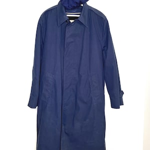 Op de afbeelding: Een marineblauwe trenchcoat met bijpassende capuchon. De jas heeft een kraag, lange mouwen en een knoopsluiting aan de voorkant. De jas hangt tegen een witte muur.