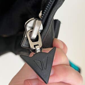 May include: Close-up of a black zipper pull with a silver zipper and a black triangle logo with the text "D.P.T."