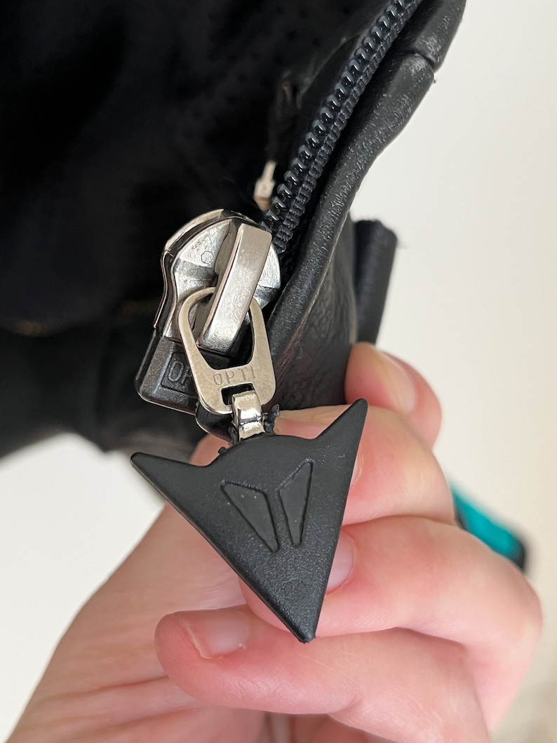 May include: Close-up of a black zipper pull with a silver zipper and a black triangle logo with the text "D.P.T."