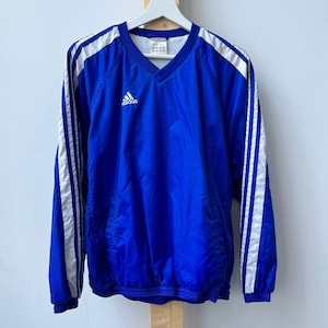 Può includere: Una giacca a vento Adidas blu con strisce bianche sulle maniche e un logo Adidas bianco sul petto.