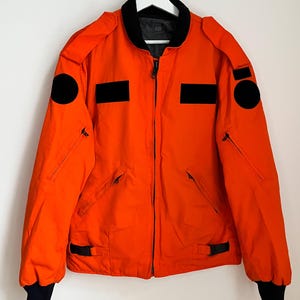 May include: Bright orange jacket with black accents, including a black collar, cuffs, and patches. Features a full-length zipper, two front pockets, and two smaller zippered pockets. The jacket is hanging on a white hanger.