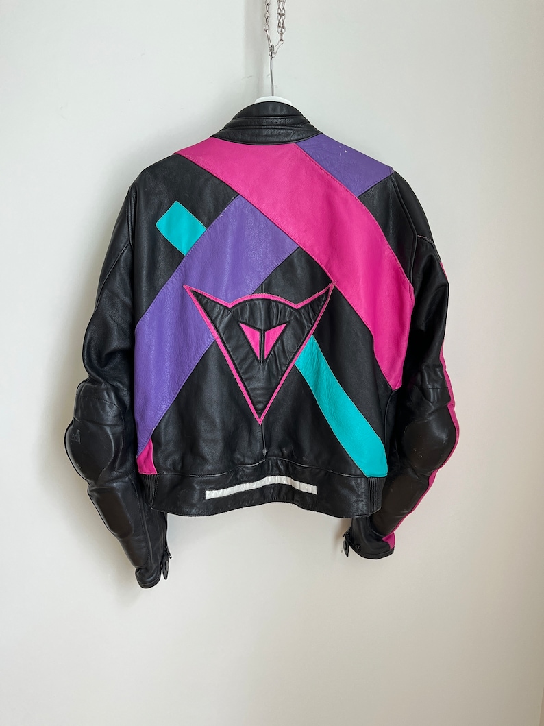 May include: Black leather motorcycle jacket with colorful geometric panels in pink, purple, and turquoise. The jacket has a large Dainese logo on the back.