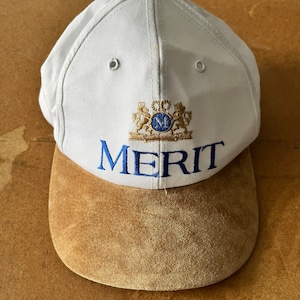 Op de afbeelding: Witte baseballpet met een bruine suède klep. De voorkant van de pet heeft een geborduurd gouden embleem met een blauwe "M" en het woord "MERIT" in blauwe borduurwerk.