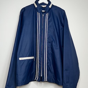 Può includere: Giacca a vento blu navy con strisce bianche sul davanti e cerniera bianca. La giacca ha un colletto bianco e una tasca con cerniera bianca sul lato sinistro. È appesa a un'appendiabiti bianco.