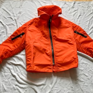 May include: A bright orange jacket with a zipper closure and black accents. The jacket has two zippered pockets on the chest and two zippered pockets on the sleeves.