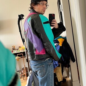 May include: A person wearing a black beanie, clear glasses, and a multi-colored leather jacket with a teal, purple, pink, and black color scheme. The person is wearing blue jeans and is standing in front of a mirror.
