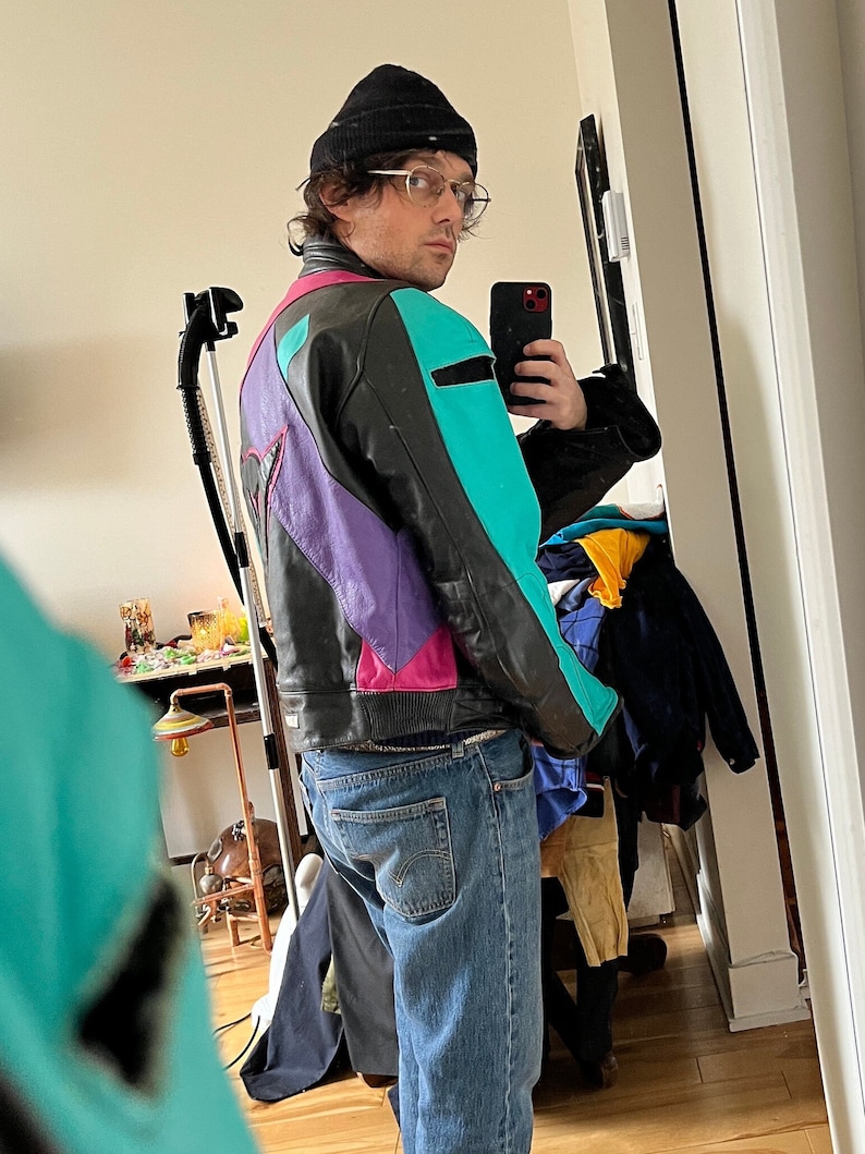 May include: A person wearing a black beanie, clear glasses, and a multi-colored leather jacket with a teal, purple, pink, and black color scheme. The person is wearing blue jeans and is standing in front of a mirror.