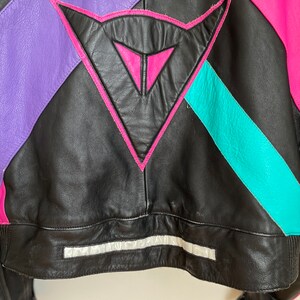 May include: Black leather jacket with pink, purple, and turquoise accents. The jacket features a large triangle logo on the back.
