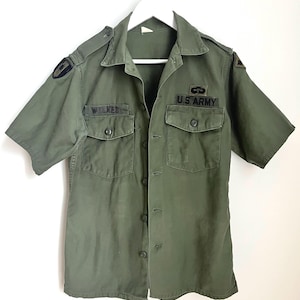 Peut inclure: Chemise militaire vert olive à manches courtes avec deux poches de poitrine et des boutons. Comprend des écussons sur les épaules et les inscriptions "WILKES" et "U.S. ARMY". La chemise est suspendue à un cintre blanc.