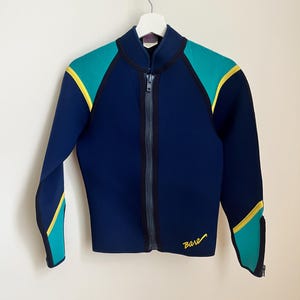 Peut inclure: Veste de combinaison de plongée bleu marine avec des touches de turquoise et de jaune. La veste est dotée d'une fermeture éclair intégrale et le mot "Bare" est écrit en jaune. Suspendue à un cintre blanc.