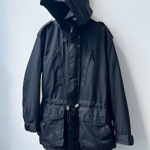 Op de afbeelding: Een zwarte capuchonjas met meerdere zakken en een trekkoord in de taille.