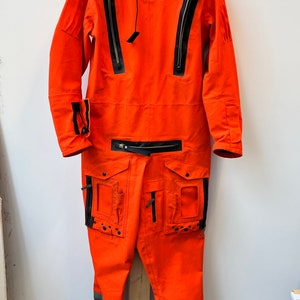 Może przedstawiać: Jas w kolorze jaskrawo pomarańczowym z czarnymi akcentami i detalami w postaci zamków błyskawicznych. Komplet posiada wiele kieszeni i wysoki kołnierz. Komplet jest zawieszony na wieszaku.