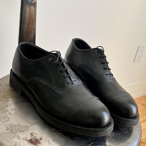 Puede incluir: Un par de zapatos negros de cuero con cordones y puntera de captoe. Los zapatos tienen una suela gruesa de goma y están sobre una silla de metal.