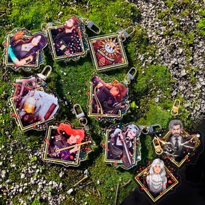 Op de afbeelding: Een verzameling van acht vierkante acryl sleutelhangers met diverse geïllustreerde personages in donkere kaders met rode bloemblaadjes. Elke sleutelhanger heeft een zilverkleurige karabijnhaak. Eén vierkant toont een zonembleem met de tekst WHERE WHOOO.