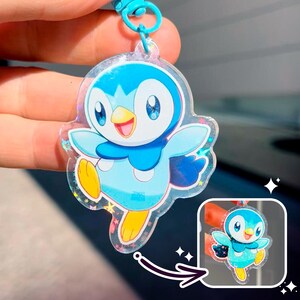 Op de afbeelding: Een heldere acryl sleutelhanger met een cartoon pinguïn karakter in tinten blauw, wit en geel. De sleutelhanger heeft een blauwe lus en een kleine, gedetailleerde afbeelding van het karakter. Het karakter lacht met uitgestrekte armen.