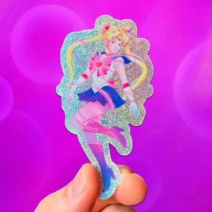 May include: A holographic sticker featuring a Sailor Moon character in a dynamic pose. The character is depicted in a sailor uniform with a pink bow, blue skirt, and pink and blue boots. The sticker has a glittery, iridescent finish and is held against a purple background.