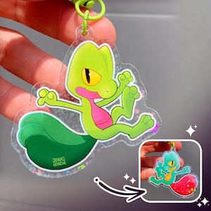 May include: A clear acrylic keyring featuring a green and pink cartoon gecko. The gecko has yellow eyes and is posed with its arms and legs outstretched. The keyring has a green loop and the artist's name, "JONAS GRANCHA", is printed on the bottom.