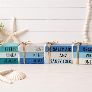 May include: Four decorative wooden blocks with beach-themed phrases: "FEELIN' KINDA BEACHY," "GONE TO THE BEACH," "SALTY AIR AND SANDY TOES," and "BEACH VIBES ONLY." Each block is painted in shades of blue and white, tied with twine.