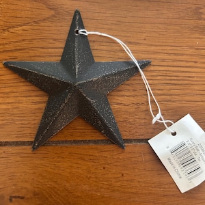 May include: A five-pointed star ornament with a dark, textured, distressed finish. A white string is threaded through a hole at the top point. A white tag attached to the string displays a barcode and the text: Rusty Star-Antique, 3.5"x3.5" dia. Tin.