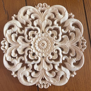 May include: A circular light-colored wooden carving featuring an intricate symmetrical floral pattern with carved petals and scrolling leaf motifs. The design is centered around a small flower and radiates outward with detailed relief work.