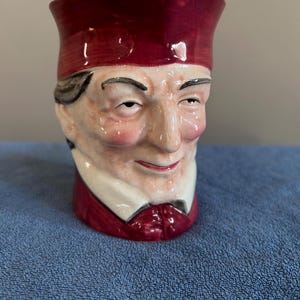 May include: A ceramic mug shaped like a man's head, featuring a burgundy hat and suit. The mug has a detailed face with rosy cheeks and a slight smile. The mug is a unique decorative item.