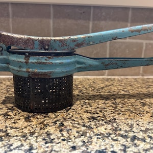 以下が含まれることがあります： ヴィンテージのポテトマッシャー。使い込まれたターコイズブルーの仕上げと黒い穴あきバスケットが特徴です。ハンドルには錆の跡が見られます。ジャガイモなどの柔らかい食品を潰すためのキッチン用品です。