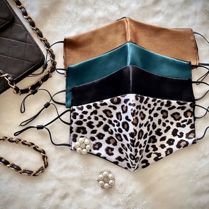 Pode incluir: Um conjunto de cinco máscaras faciais, incluindo uma máscara de estampa de leopardo, uma máscara preta, uma máscara turquesa e duas máscaras marrons. As máscaras estão empilhadas, com a máscara de estampa de leopardo no topo. Todas as máscaras são feitas de seda e têm um toque suave e luxuoso.