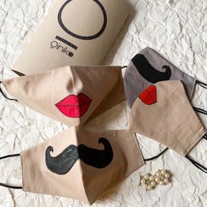 Puede incluir: Cuatro mascarillas de algodón beige con diferentes diseños. Dos mascarillas tienen bigotes negros, una tiene un labio rojo y otra tiene un bigote negro y un labio rojo. Las mascarillas están sobre una superficie blanca texturizada. El texto "QINIKO" está en una caja de papel marrón.