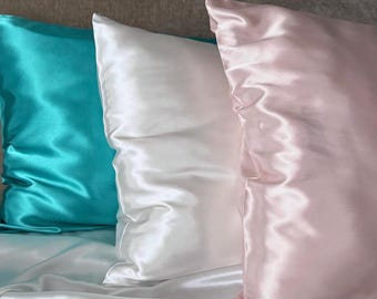 May include: Three satin pillowcases in different colors: turquoise, white, and pink. The pillowcases are arranged on a white satin bed sheet.