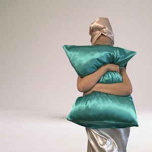 May include: A person wearing a light pink satin hair wrap holds a teal satin pillow in front of their face.