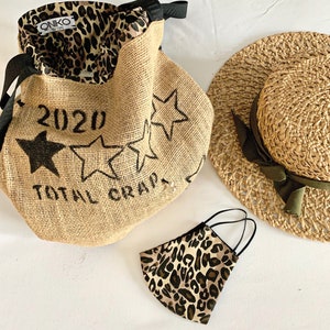 May include: A brown burlap tote bag with black star designs and the text "2020 Total Crap". The bag has a leopard print lining and a brown straw hat with a green ribbon is in the background. A leopard print face mask is in the foreground.