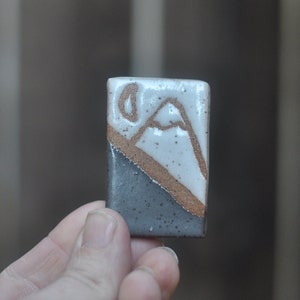 May include: A small ceramic tile with a white background and a brown and black mountain design.