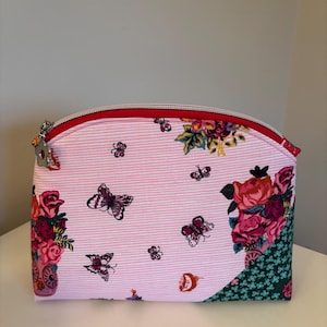 May include: Pink and white striped cosmetic bag with a red zipper and floral and butterfly patterns. The bag features a curved top and a small charm on the zipper pull. The bag has a floral pattern on the sides.