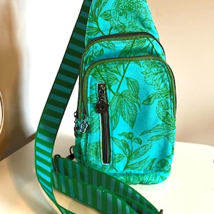 May include: A teal sling bag with a green floral pattern and green trim. The bag has three zippered compartments and a striped green and blue strap. The strap is adjustable and has a metal buckle. The bag is made of fabric.