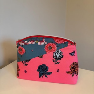 May include: A rectangular zippered pouch with a split design. The top half is teal with a floral and village pattern, and the bottom half is bright pink with rose illustrations. The zipper is red with a white pull.