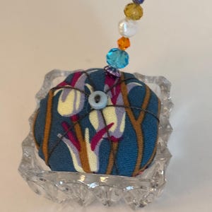 May include: A handmade pin cushion with a floral pattern in blue, yellow, and purple, topped with a beaded embellishment. The pin cushion is set in a clear, faceted glass dish. The beads are various colors.