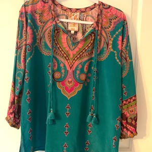 May include: A teal-colored long-sleeve blouse with a vibrant paisley pattern in pink, purple, and green. The blouse features a V-neck with tassel ties and is hanging on a white hanger. The sleeves have a patterned cuff.