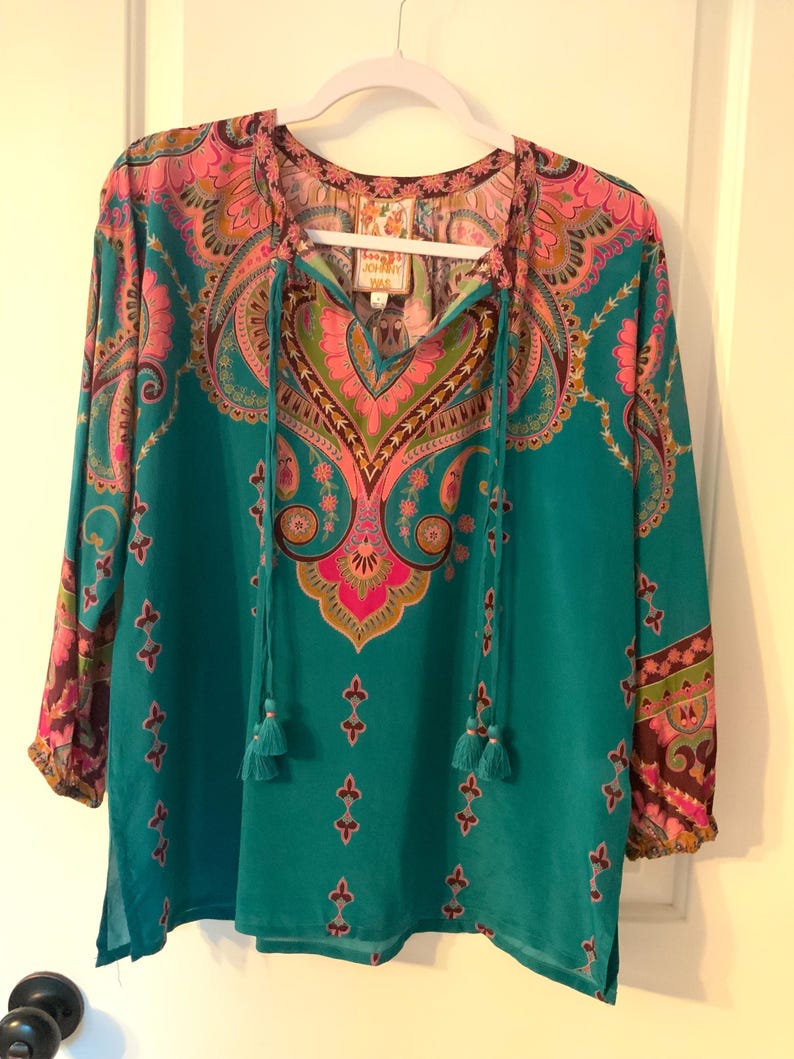May include: A teal-colored long-sleeve blouse with a vibrant paisley pattern in pink, purple, and green. The blouse features a V-neck with tassel ties and is hanging on a white hanger. The sleeves have a patterned cuff.
