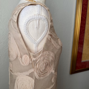 May include: A sleeveless, beige dress with a floral pattern, displayed on a mannequin. The dress features raised, textured rose-like embellishments. A gold chain necklace is draped on the mannequin's shoulder.