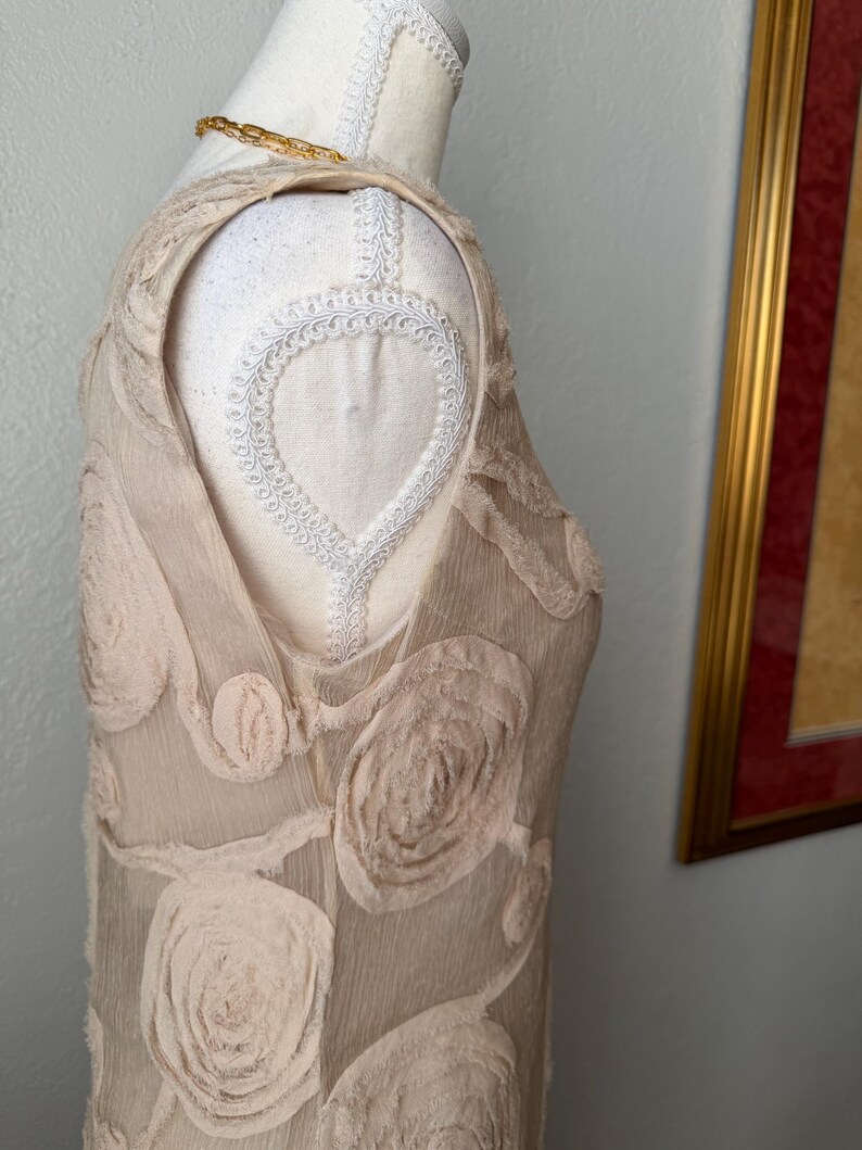 May include: A sleeveless, beige dress with a floral pattern, displayed on a mannequin. The dress features raised, textured rose-like embellishments. A gold chain necklace is draped on the mannequin's shoulder.
