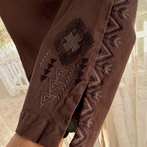 May include: Close-up of a brown garment sleeve with detailed embroidery in brown and grey. The embroidery features geometric and floral patterns. The sleeve is buttoned at the cuff.