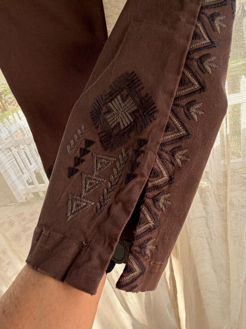 May include: Close-up of a brown garment sleeve with detailed embroidery in brown and grey. The embroidery features geometric and floral patterns. The sleeve is buttoned at the cuff.