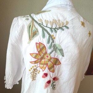 May include: White blouse with floral embroidery on the back. The embroidery features a large flower with yellow, orange, and brown petals, along with green leaves and red accents. The blouse has a collared neckline and short sleeves.